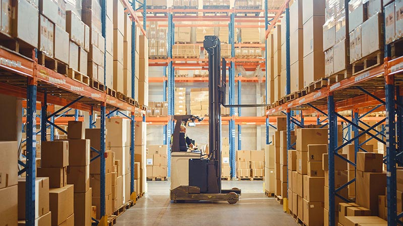 Retail warehouse full of shelves with goods: Electric Forklift Truck operator lifting pallet with cardboard box on a shelf. Working in logistics storehouse product logistics and delivery center.
